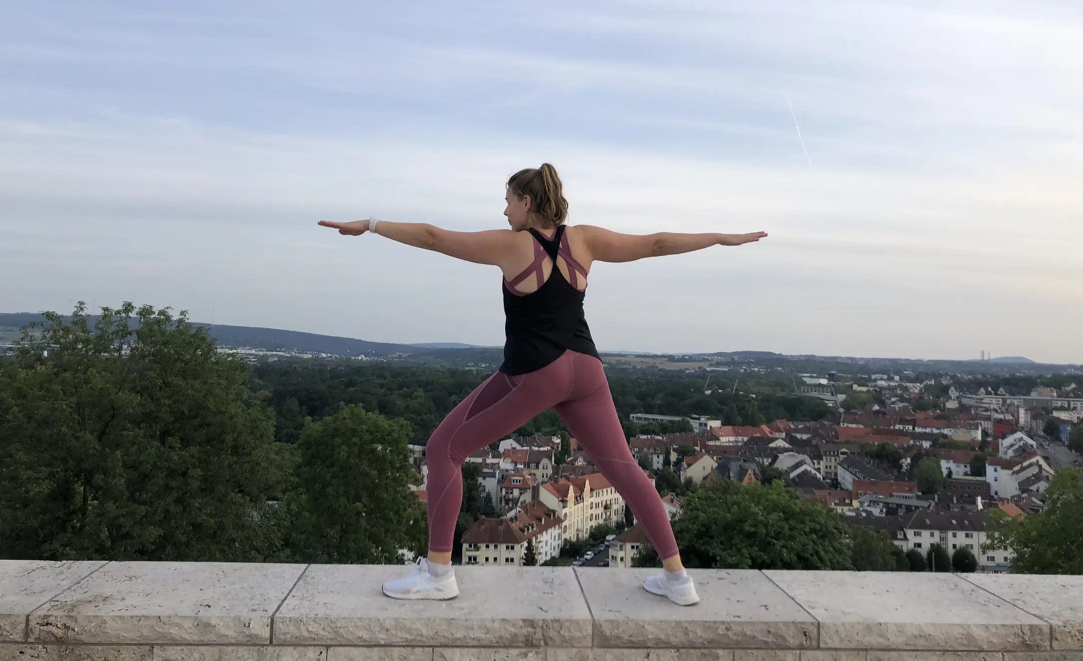 Annkathrin steht in Sportsachen der Yoga-Pose Warrior auf einer Mauer. Hinter ihr ist die Skyline von Kassel.
