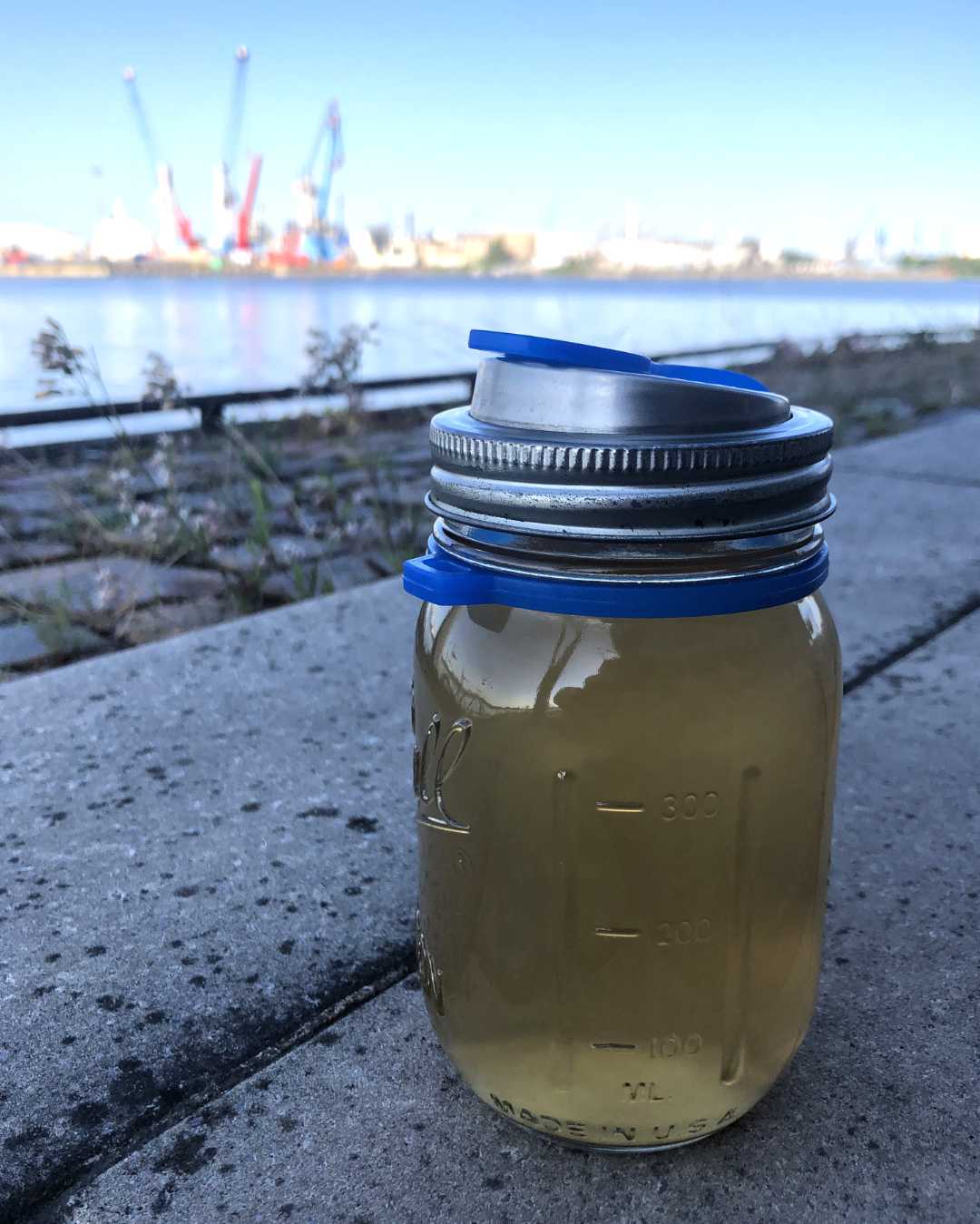 Ein Mason Jar mit dem Trinkdeckel von ecojarz steht auf Steinen. Im Hintergrund ist schemenhaft der Hamburger Hafen zu erkennen.