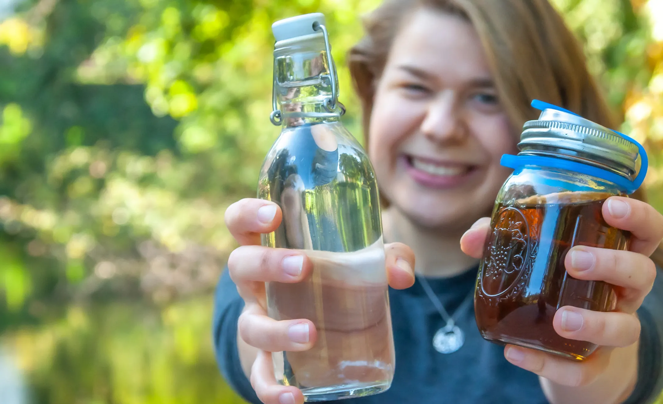 Annkathrin hält eine Glasflasche mit Wasser und einen To-Go-Becher in die Kamera. Im Hintergrund lacht sie.