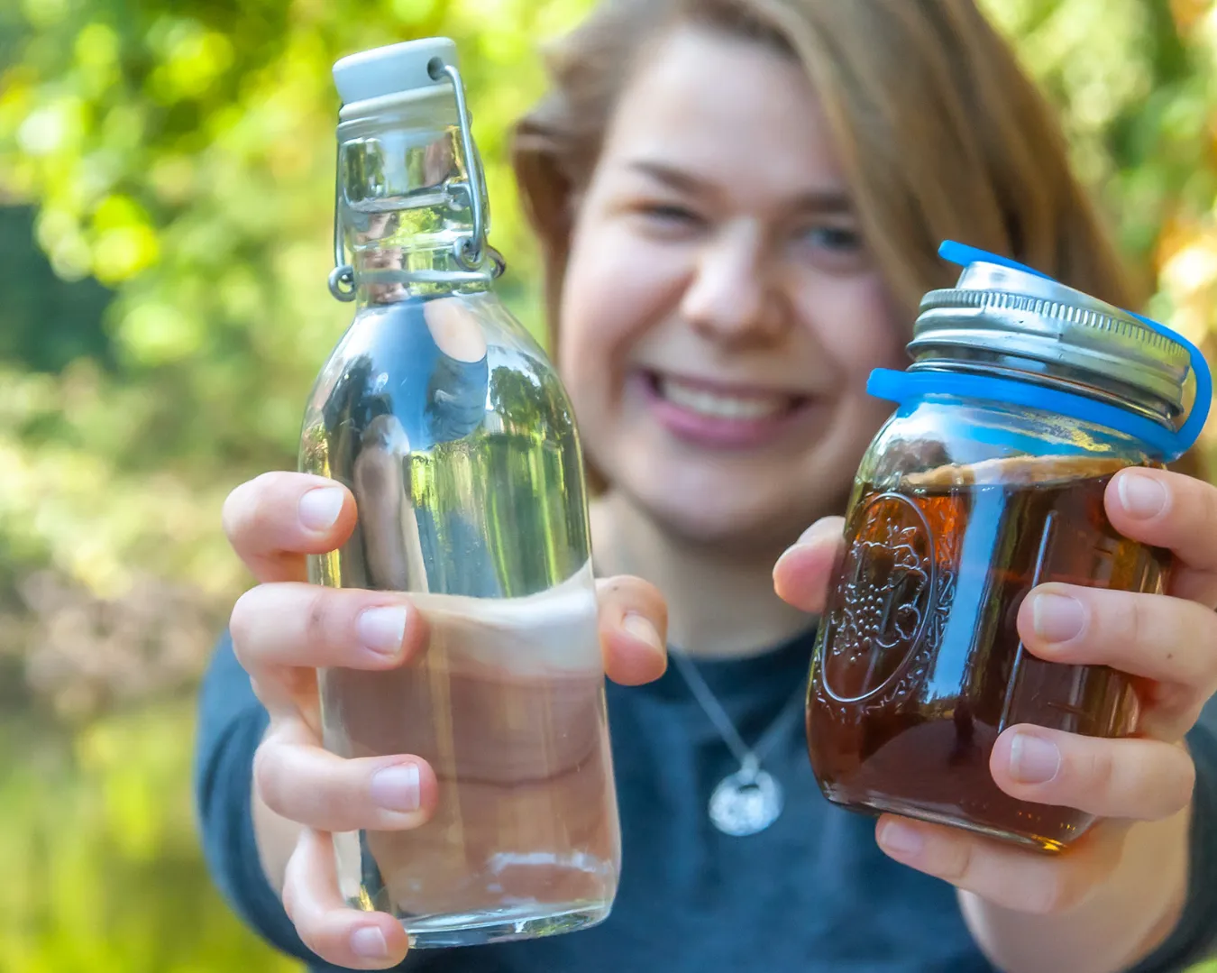 Annkathrin hält eine Glasflasche mit Wasser und einen To-Go-Becher in die Kamera. Im Hintergrund lacht sie.