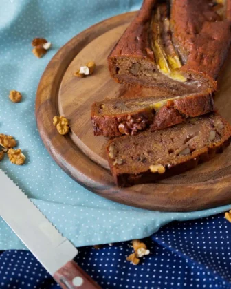 Ein aufgeschnittenes Bananenbrot auf einem Holzteller. Daneben liegen ein Brotmesser und Walnusskerne.