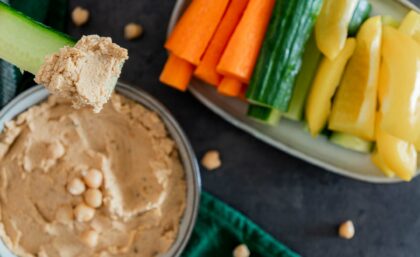 Blick aus der Vogelperspektive auf eine Schüssel mit Hummus. Daneben Gemüsesticks.