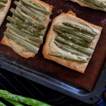 auf einem Backblech liegen fertige Spargel-Tartletts: Blätterteig-Quadrate, auf dem vegane Creme Fraîche und grüner Spargel ist.