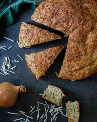 Veganes Zwiebelbrot, dass angeschnitten ist. Daneben liegt zur Deko eine Zwiebel und Käse.