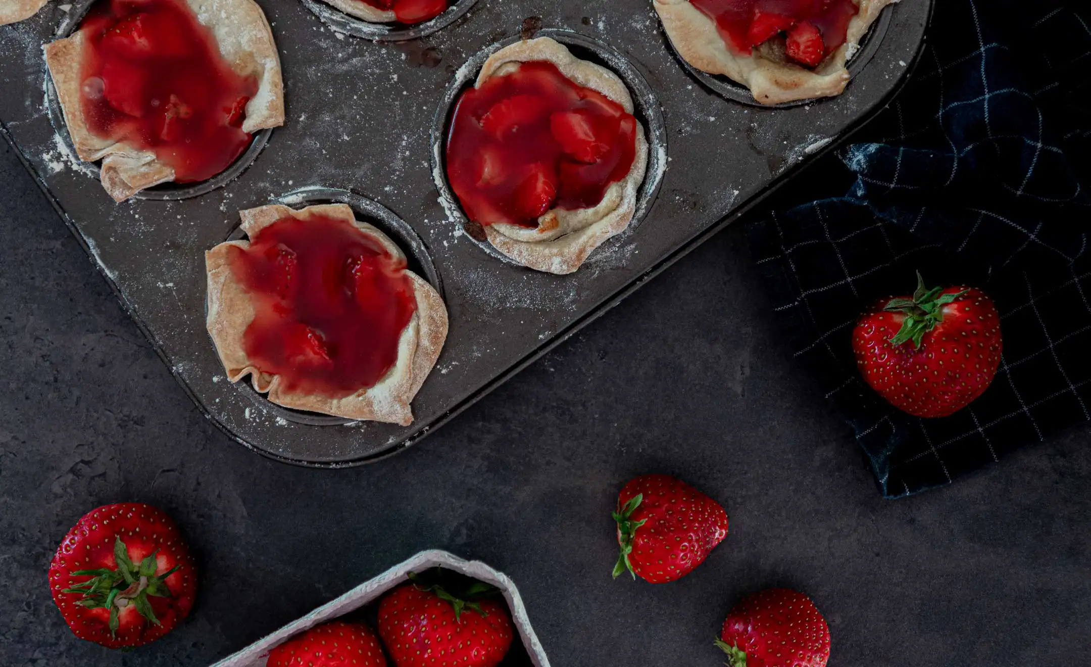 In einer Muffinblech-Backform sind Erdbeertörtchen von oben fotografiert. Daneben liegen zur Deko ein paar Erdbeeren.