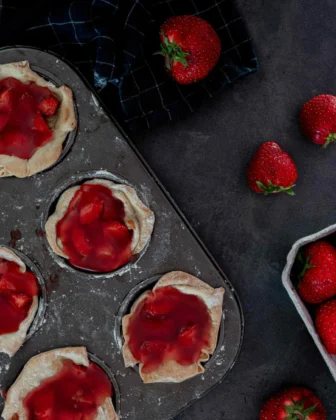 In einer Muffinblech-Backform sind Erdbeertörtchen von oben fotografiert. Daneben liegen zur Deko ein paar Erdbeeren.