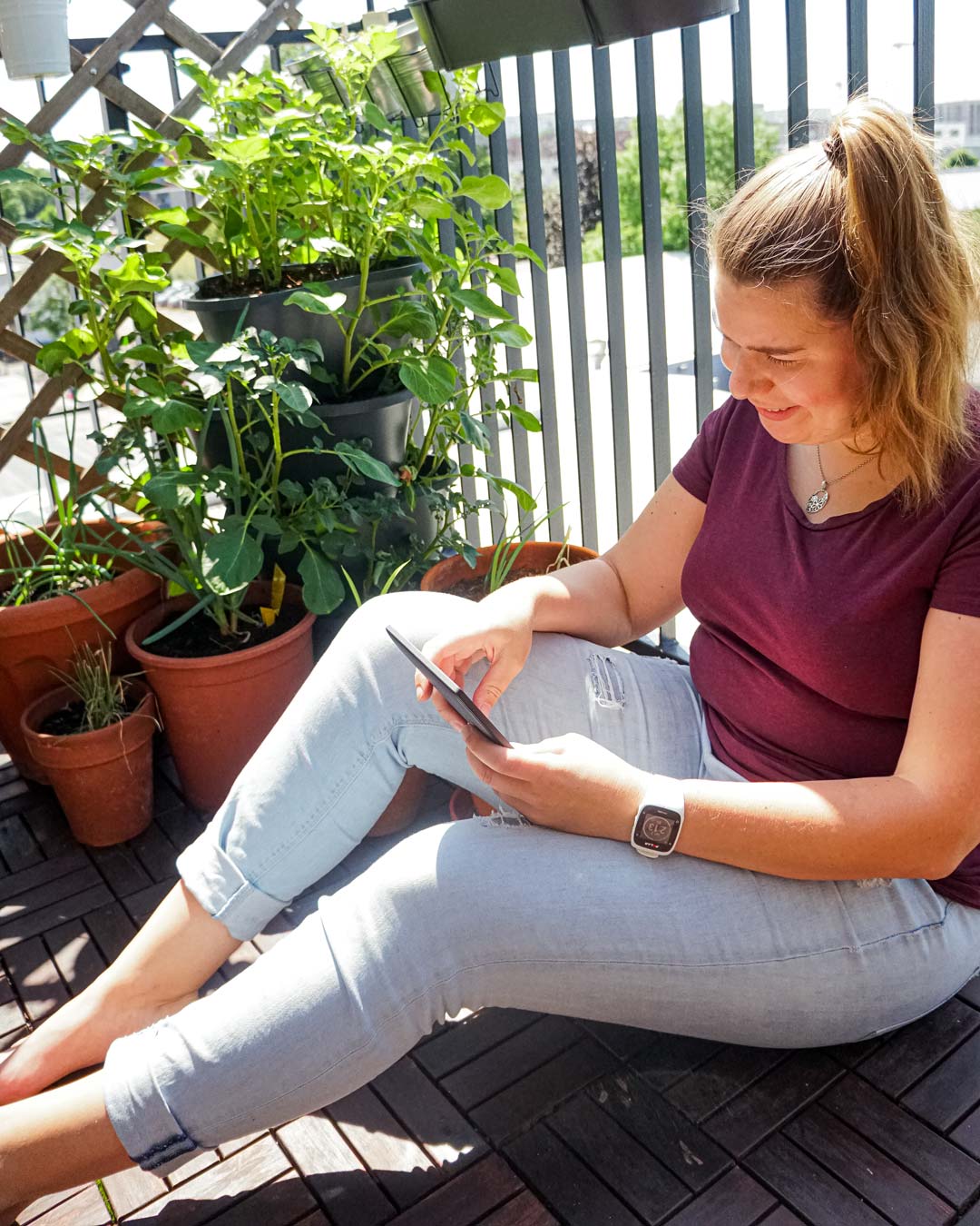 Annkathrin sitzt auf dem Balkon im T-Shirt und Jeans und liest lächelnd auf ihrem eReader. Die Sonne scheint, hinter ihr ist ihr Balkongarten zu sehen.