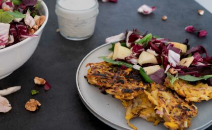 Auf einem Teller liegen vegane Kürbis-Rösti, daneben Radicchio-Salat. Im Hintergrund ein Becherchen mit Sauce.