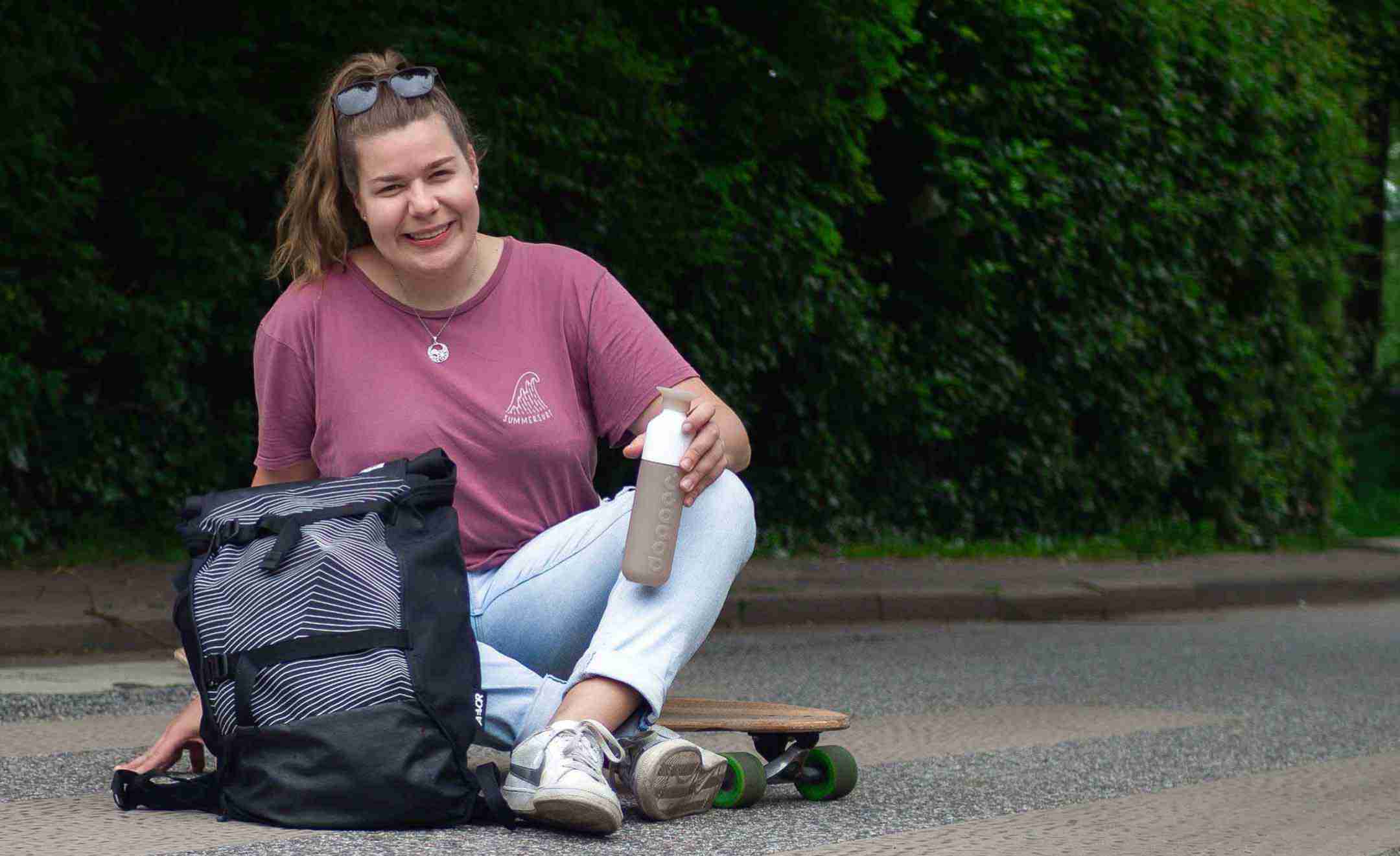 Annkathrin sitzt auf einem Longboard und lacht in die Kamera. Neben ihr steht ein Rucksack und sie hält eine Trinkflasche in der Hand.