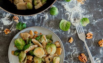 Gnocchi-Rosenkohl-Pfanne auf einem Teller drapiert, daneben Walnüsse, Rosenkohlblätter und eine Gabel.