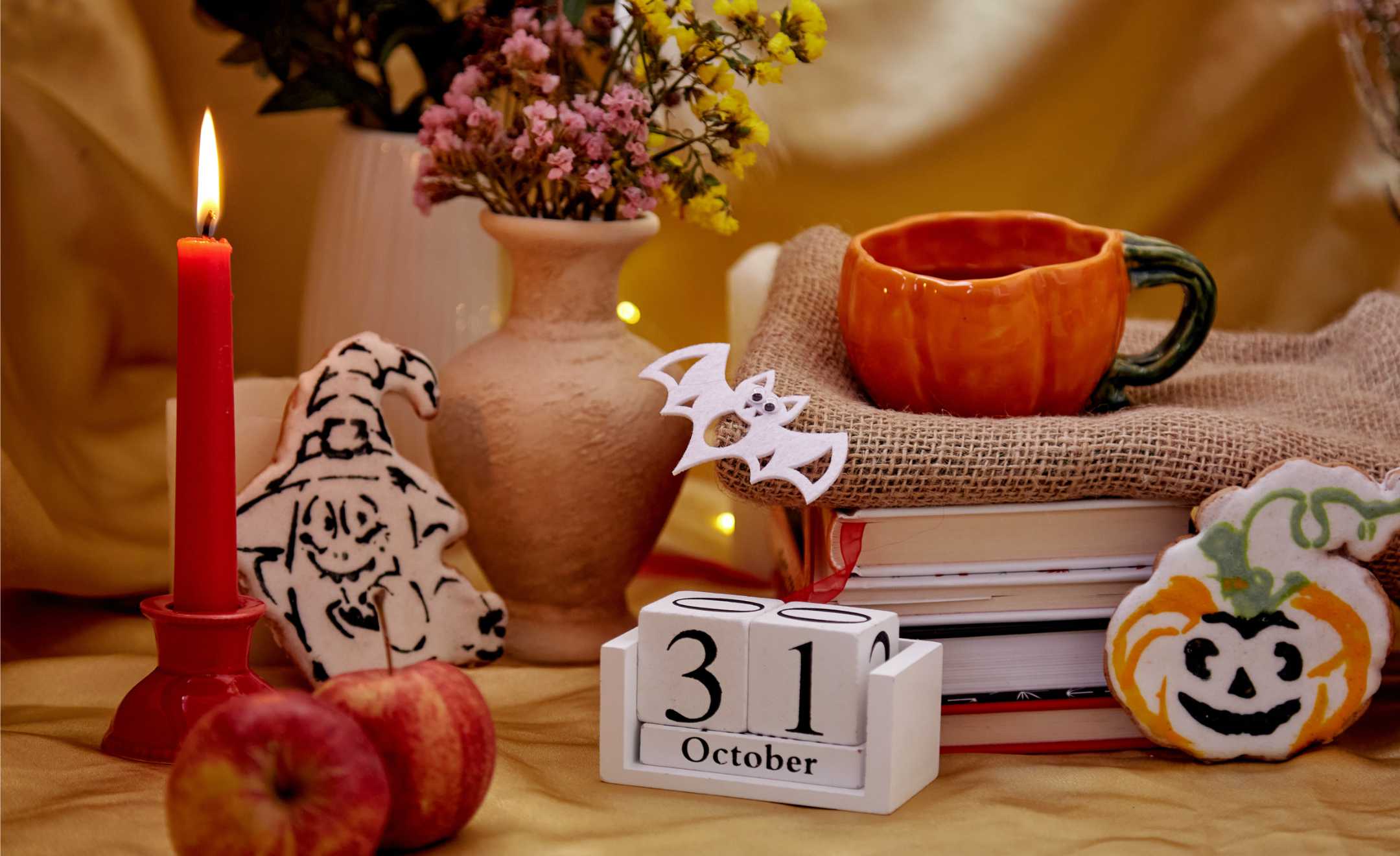 viele Halloween-Dekoartikel zusammengestellt, brennende Kerze, Kürbis-Tasse mit Tee, Blumen im Terrakotta-Topf, Kekse als Kürbis und Hexe, Fledermaus aus Filz ausgeschnitten, Holzblock-Kalender mit 31 Oktober