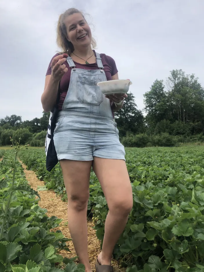 Annkathrin steht lachend in einem Erdbeerfeld. In der einen Hand hat sie eine Erdbeere, in der anderen eine Schüssel, gefüllt mit gepflückten Erdbeeren.