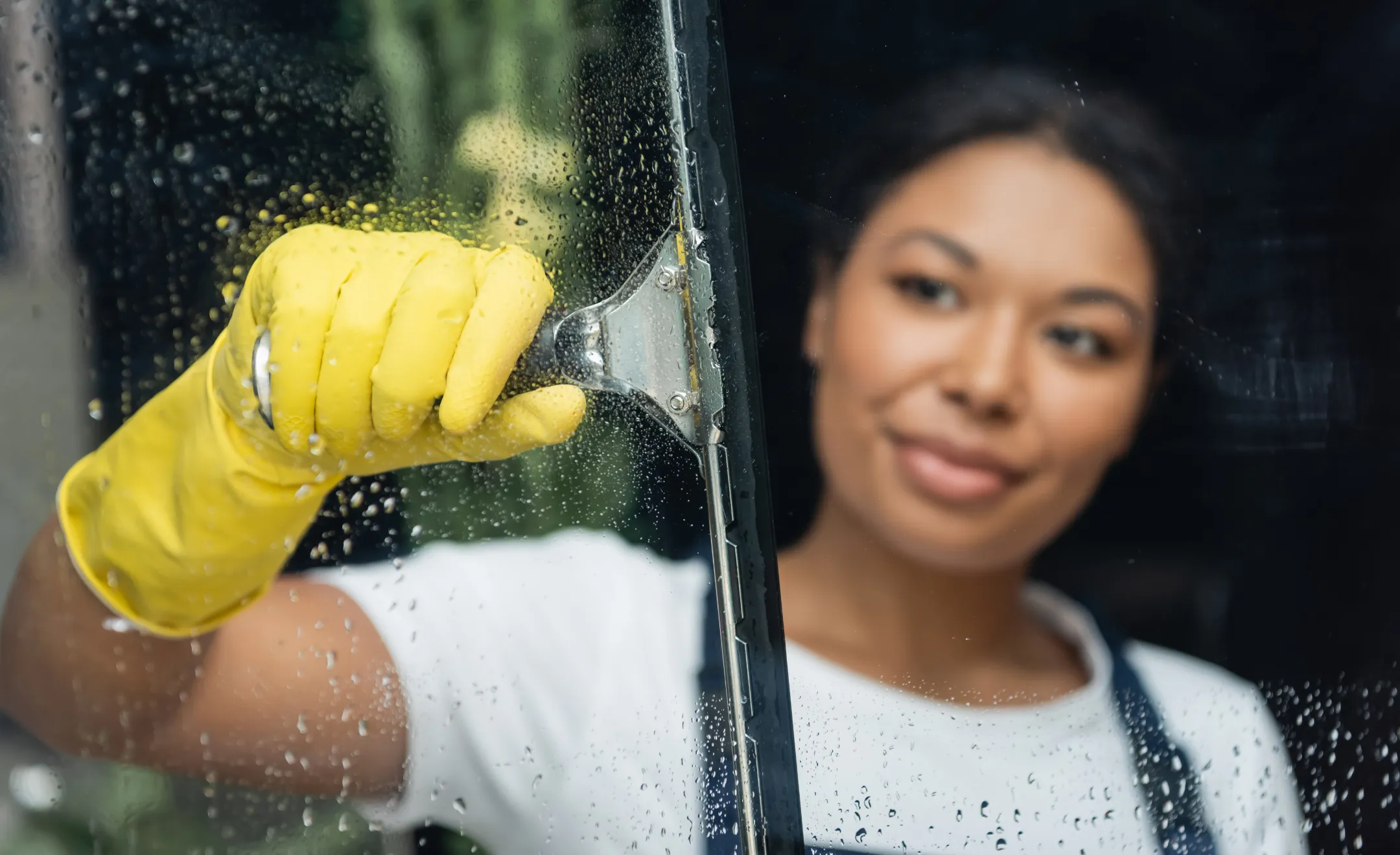 Eine Frau steht hinter einer Fensterscheibe und zieht diese mit einem Fensterabzieher ab. Die rechte Seite ist bereits sauber, auf der linken Seite des Fensters ist noch Putzmittel auf der Scheibe zu erkennen.